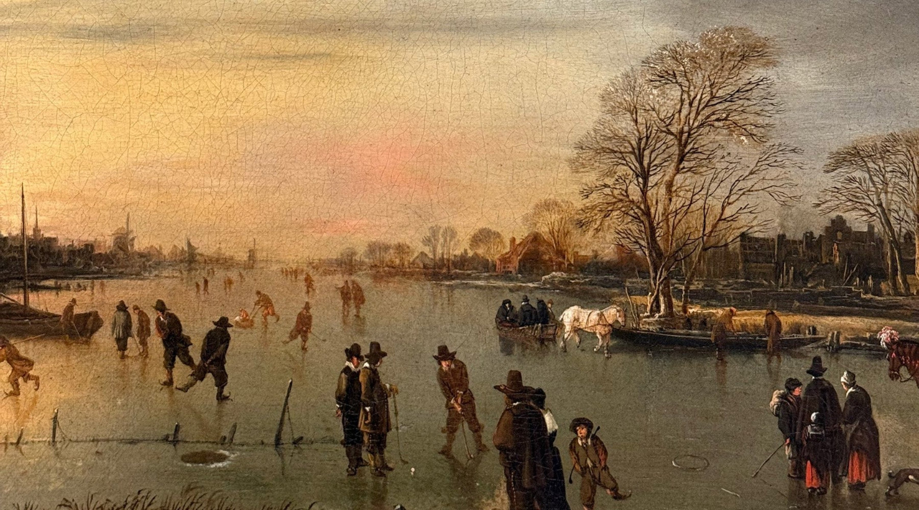 Winter scene with people skating on a frozen lake, trees, and buildings in the background.